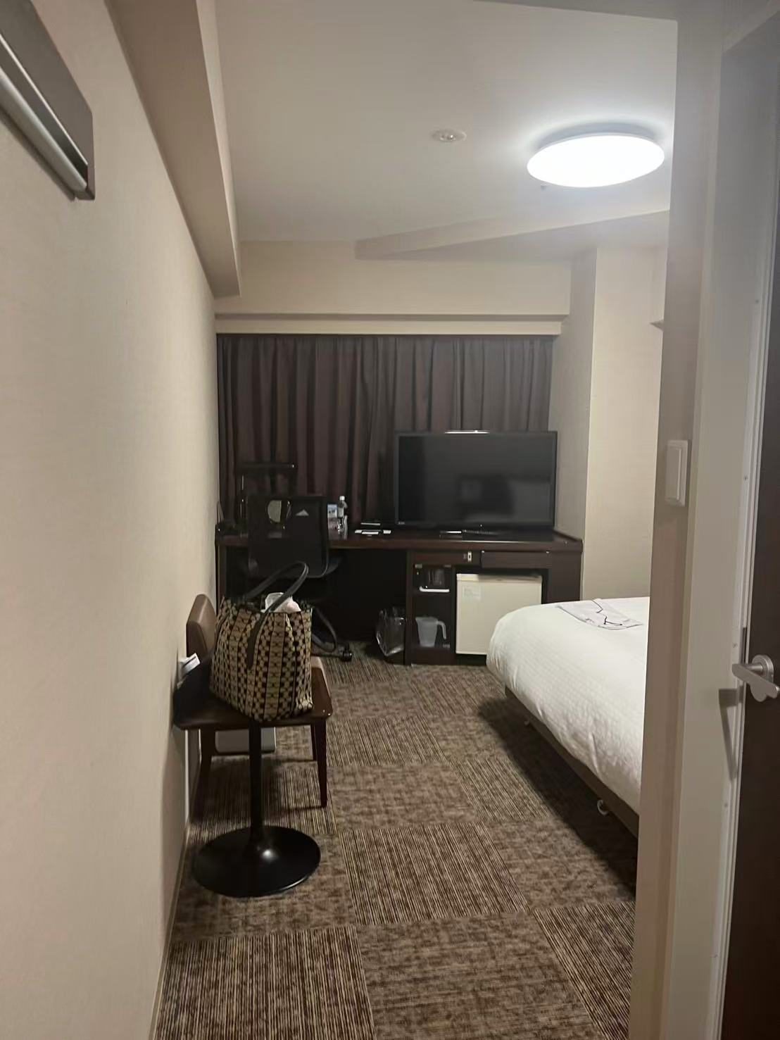 A small business hotel room seen from the door, with a tote bag on a small chair beside the door and a desk with a monitor across the room.