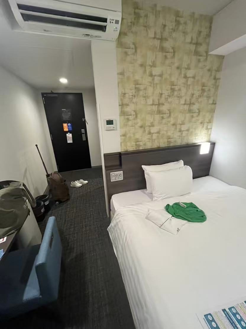 A small business hotel room seen from the door, with a wheeled bag standing upright near the entrance, shoes beside it, and the bed filling the right half of the frame.