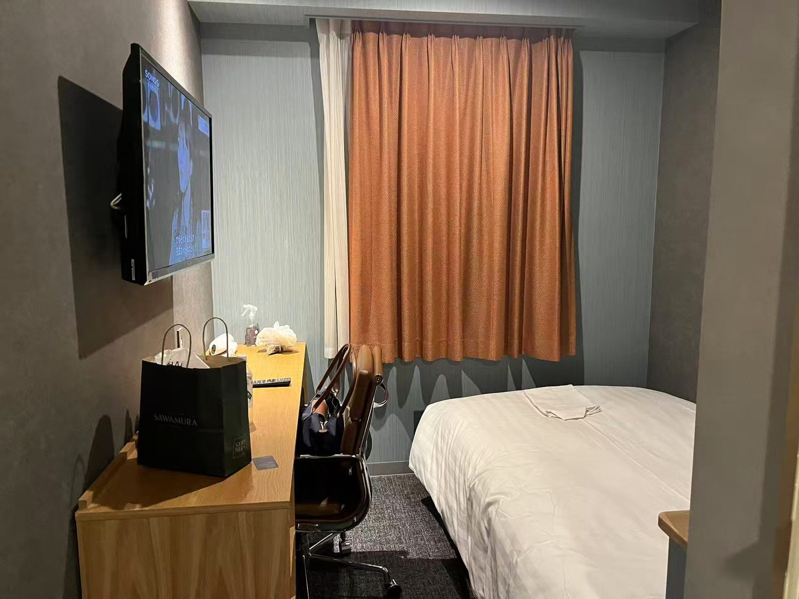 A compact business hotel room with a bed, a wall-mounted television, a desk and chair, and a curtained window.