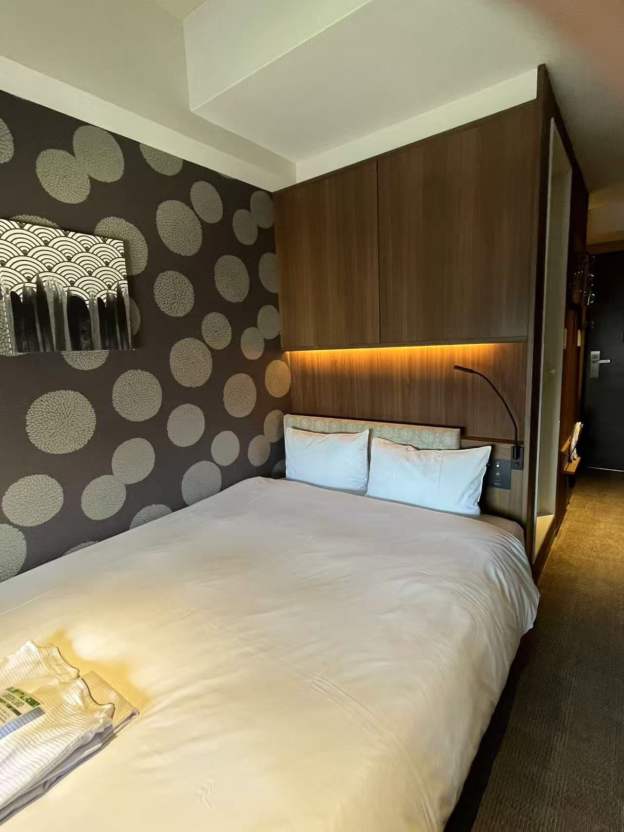 A business hotel room with a single wide bed against a patterned wall, a headboard shelf and a narrow walkway beside it.