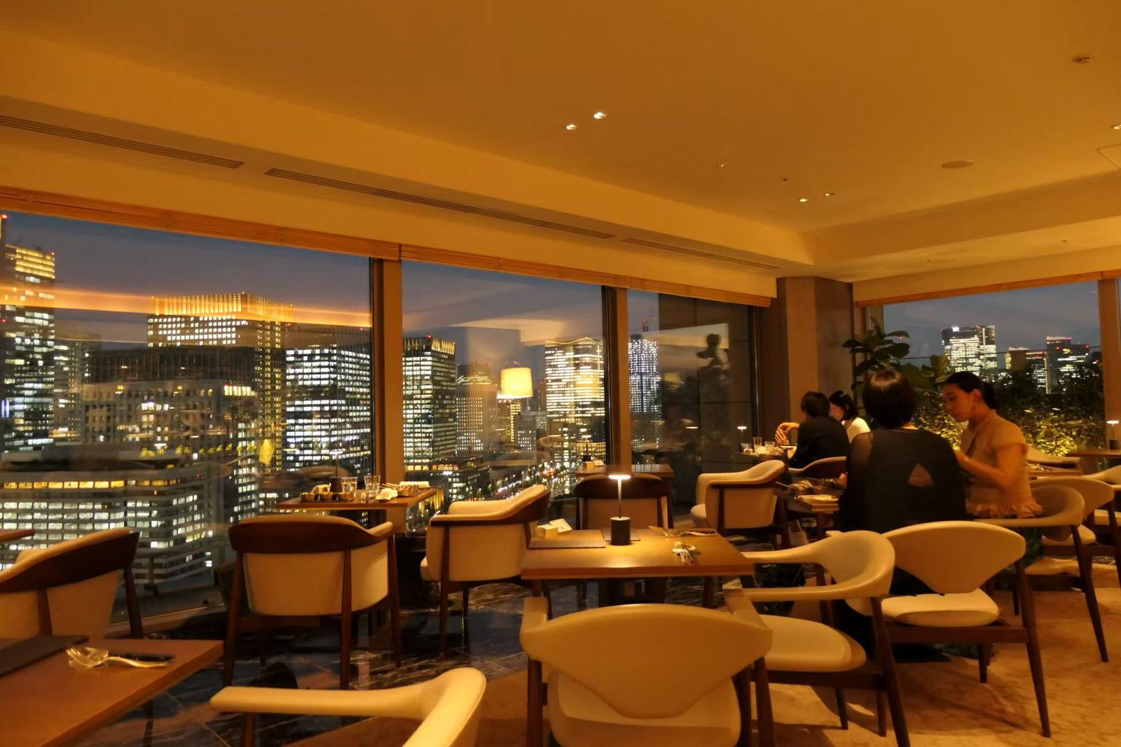 A hotel club lounge at night, with low chairs and tables along a wall of windows over a lit city.