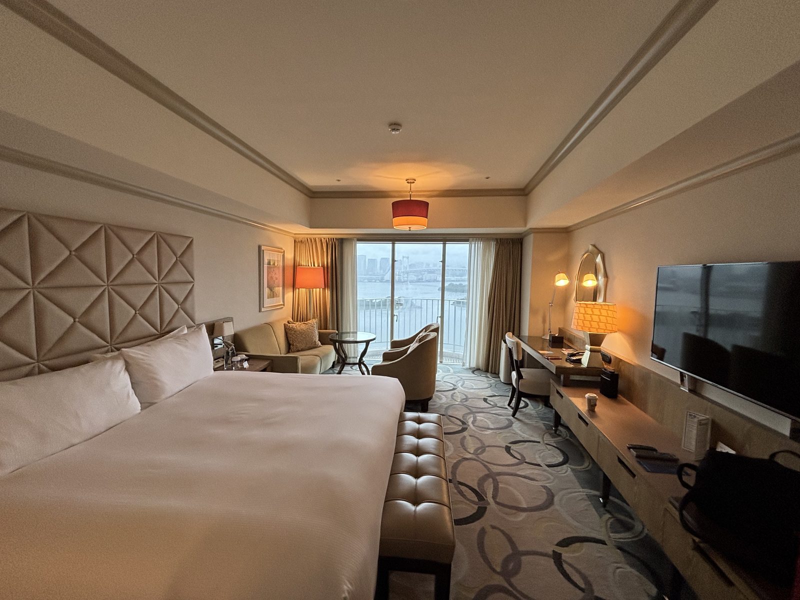 A large international-brand hotel room in Japan with a king bed, a sofa, a desk and a window onto a bay.