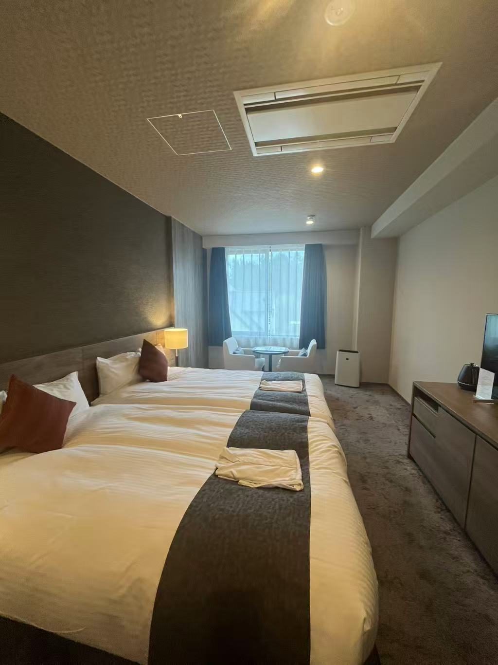 A hotel room with two single beds pushed together under one runner, a desk along the far wall and a window beyond.