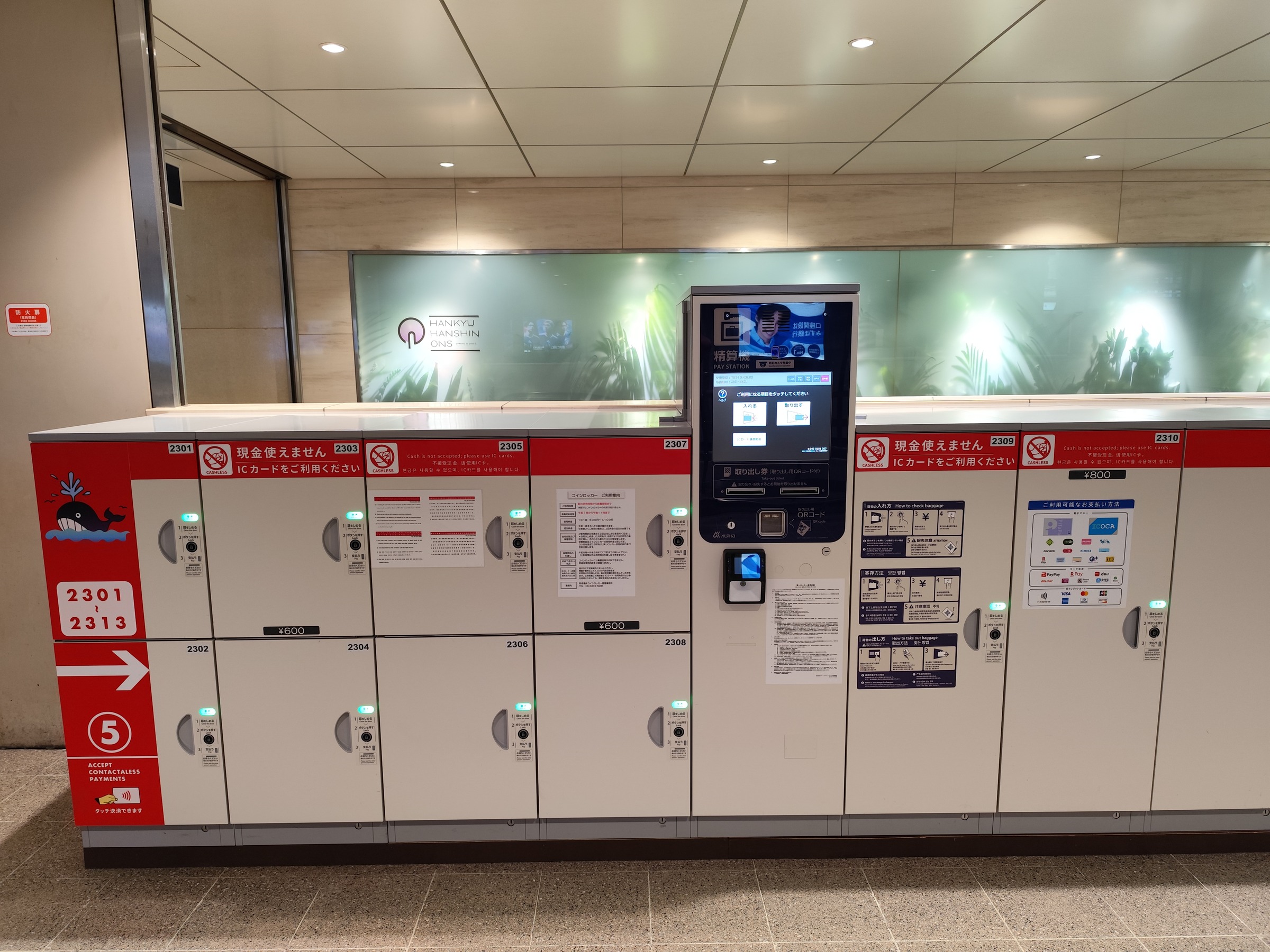 A bank of white coin lockers with red headers and a payment machine in a Japanese station.