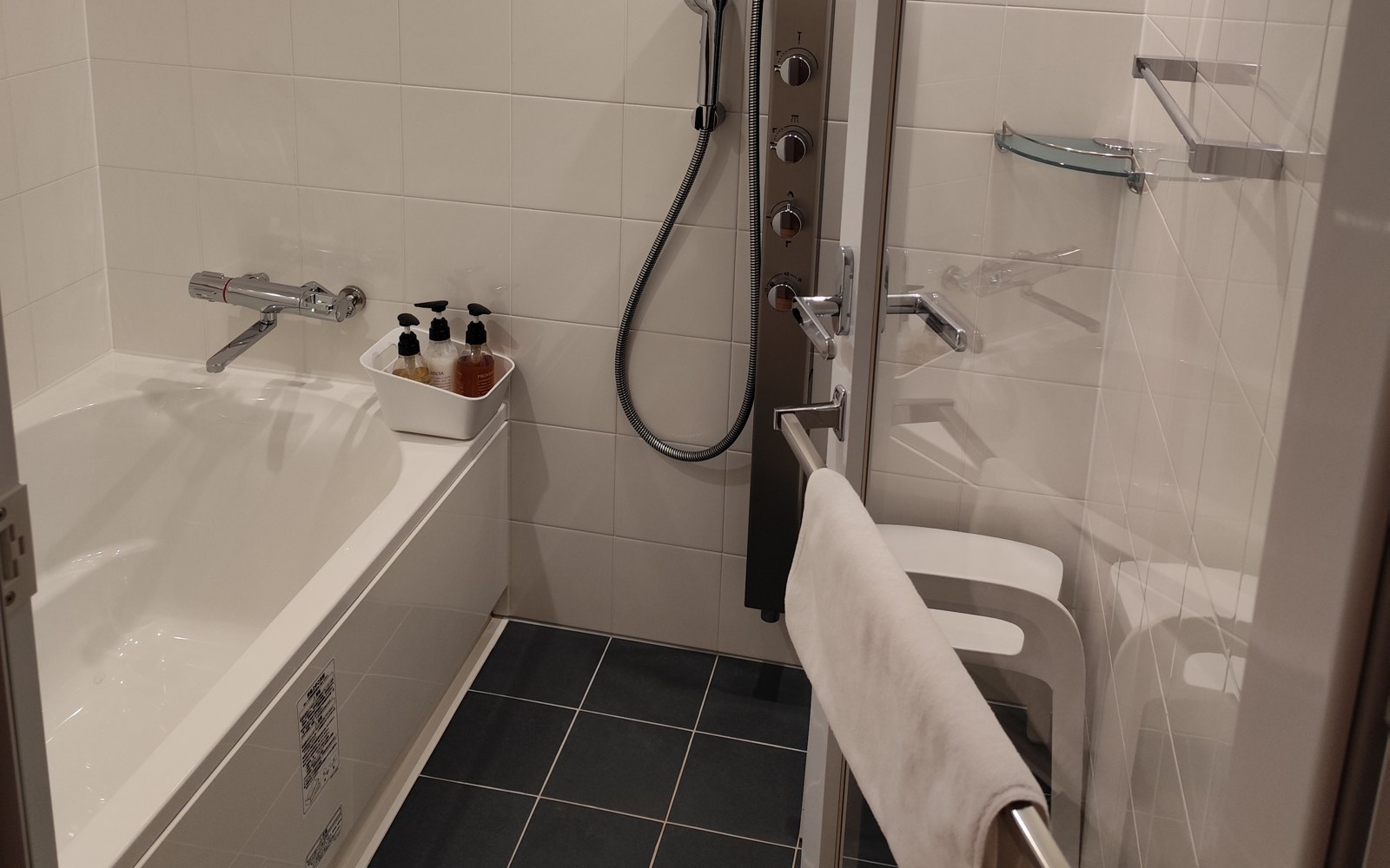 A hotel bathroom in Kyoto with a white tub on the left, a handheld shower on a metal panel, a bath stool and dark floor tiles.