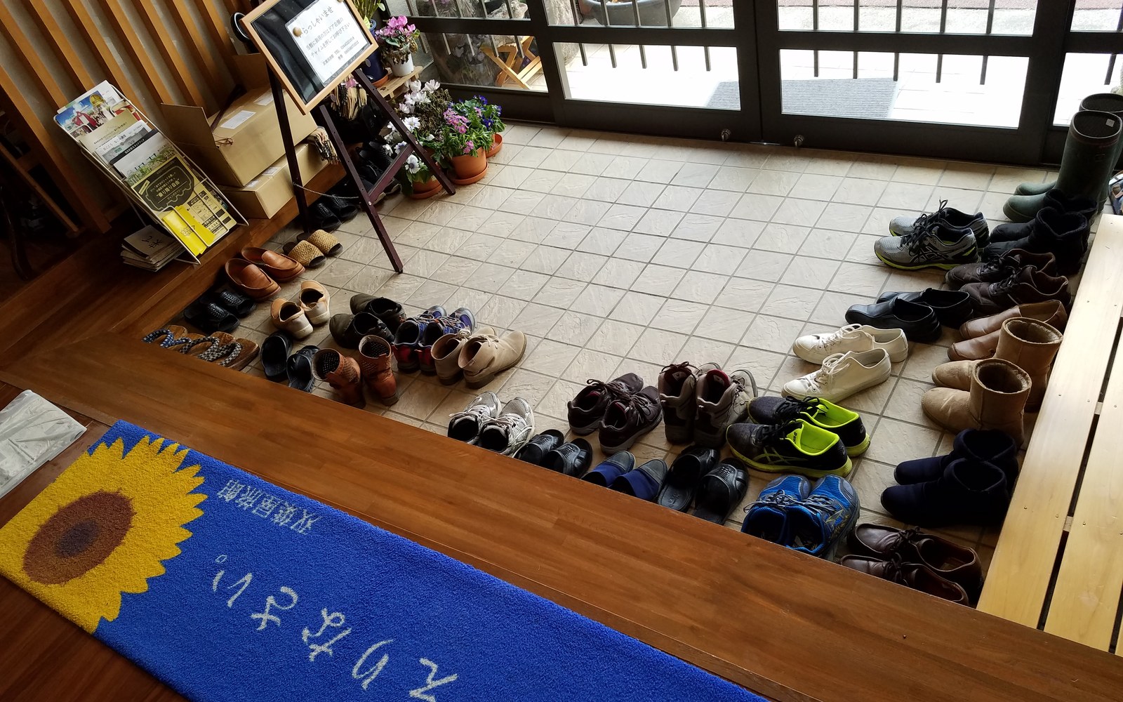 The genkan entrance of a ryokan seen from above, with many pairs of guests shoes on the tile floor and a blue welcome mat with a sunflower in the foreground.