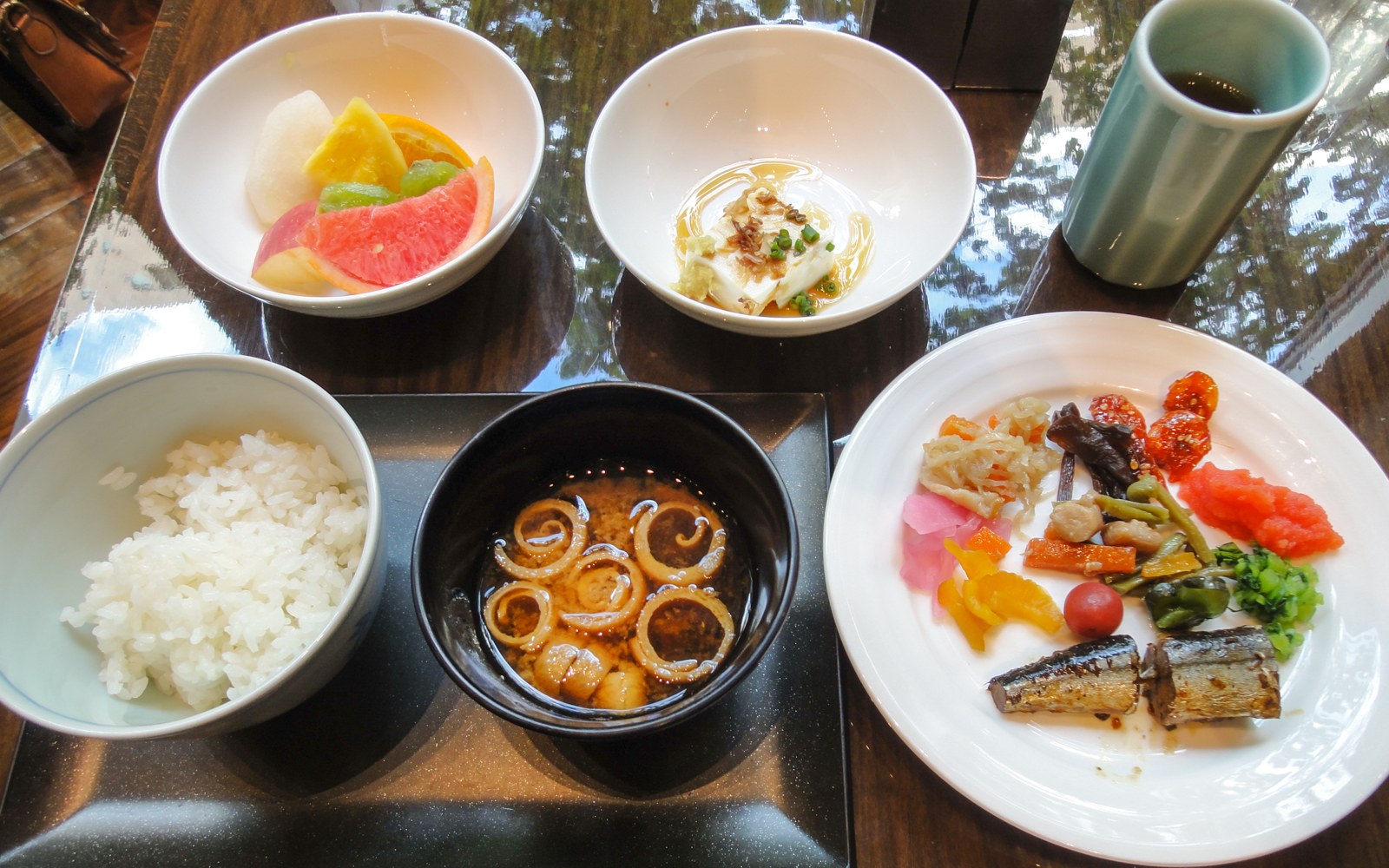 A Japanese-style hotel breakfast: a bowl of rice, miso soup, tofu, fruit, a plate of pickles and grilled fish, and a cup of tea.