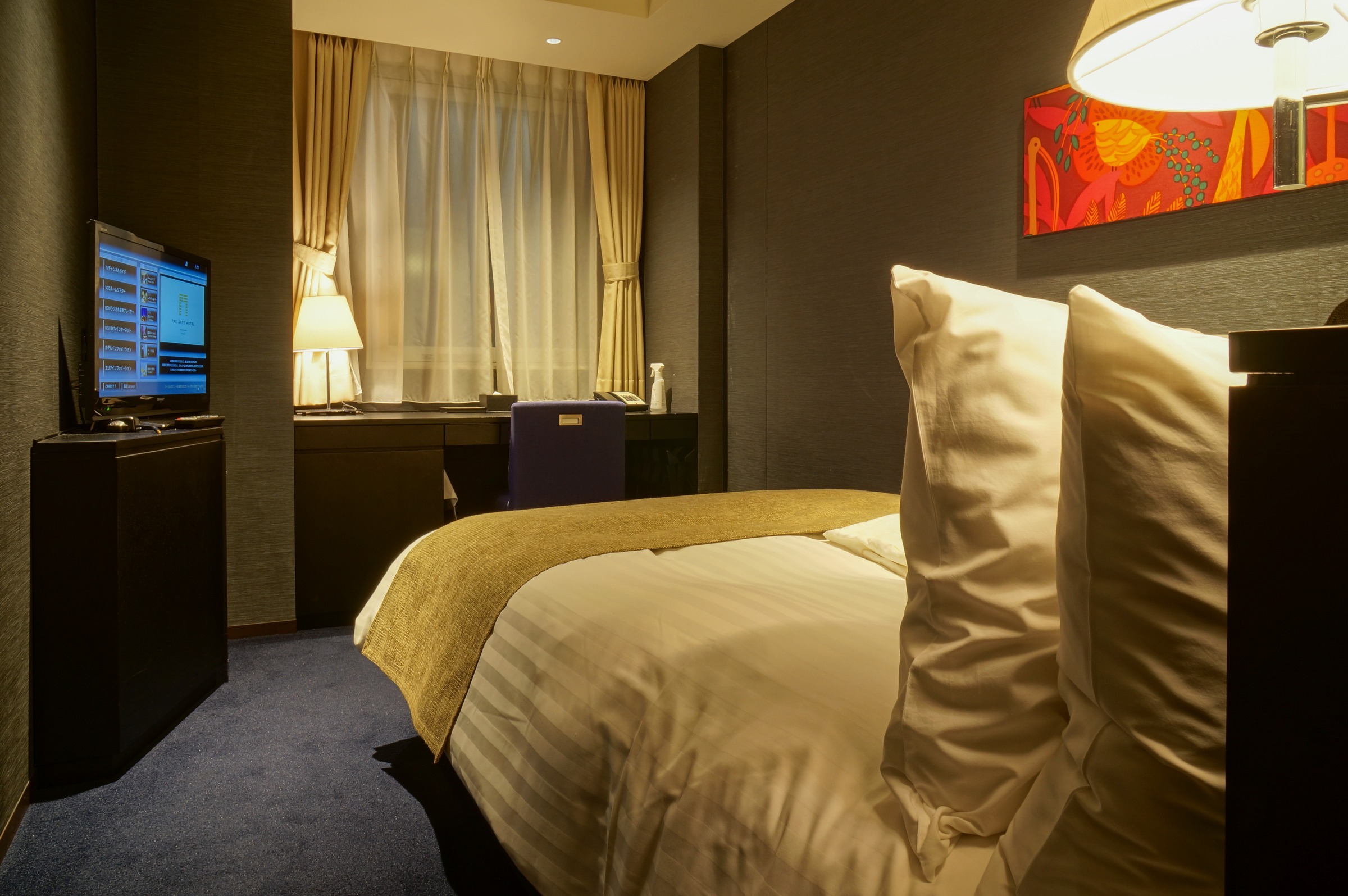 A narrow hotel room with a semi-double bed, a TV and a desk by the window.