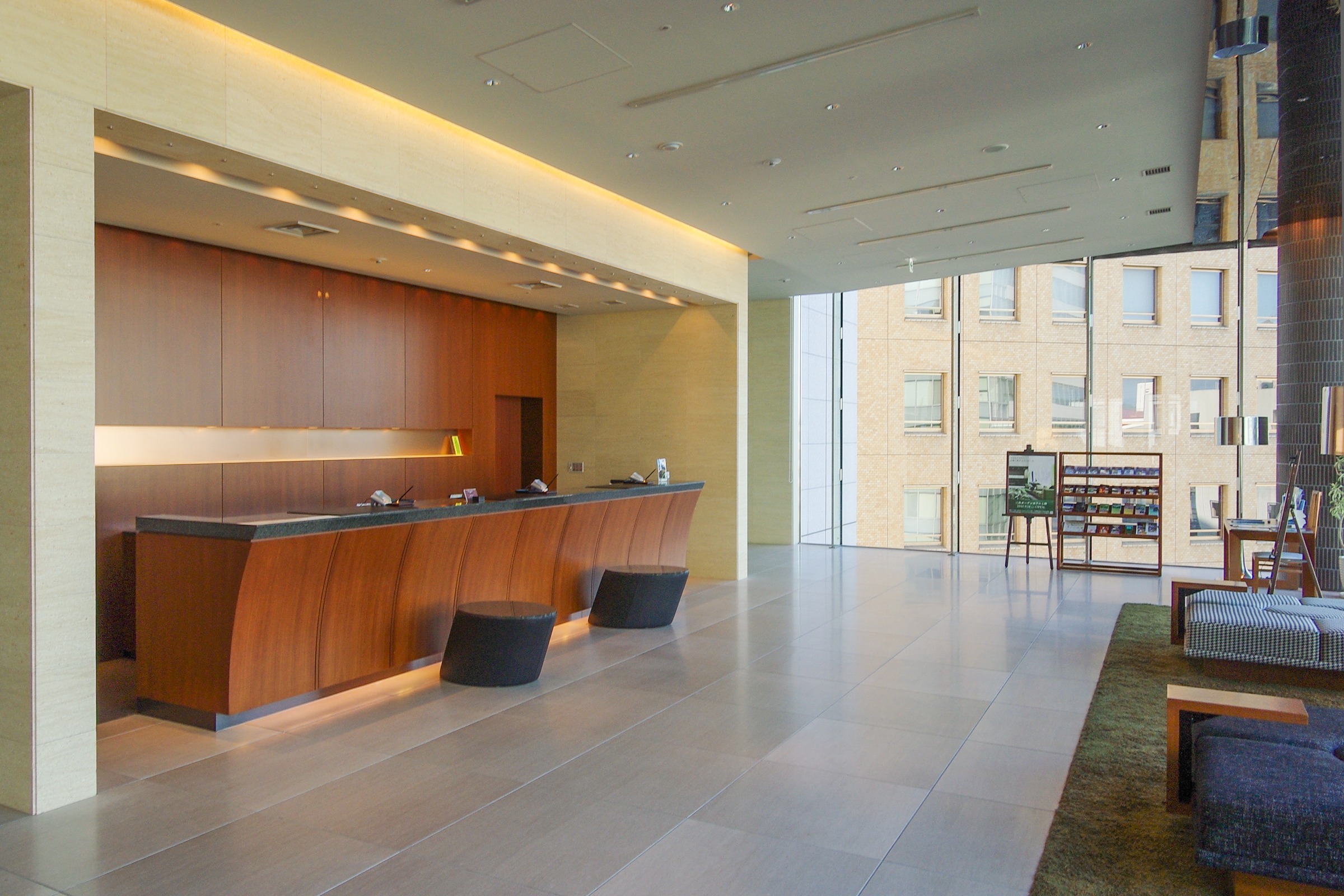 An empty hotel lobby with a long wooden front desk and a seating area by a glass wall.
