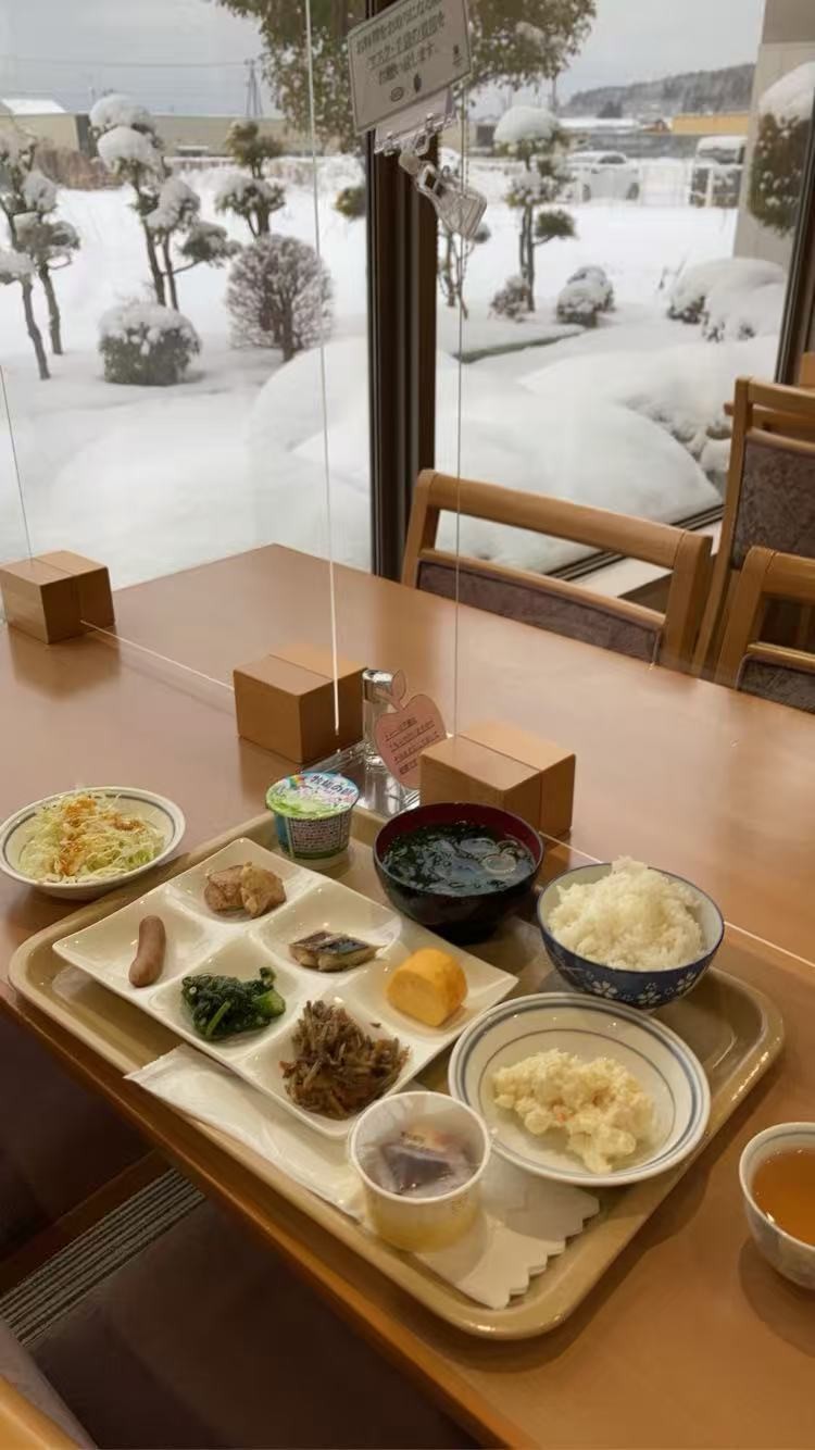 A hotel breakfast on a tray by a window with snow outside: rice, miso soup, grilled fish, spinach, a sausage, rolled egg, salad and potato salad.